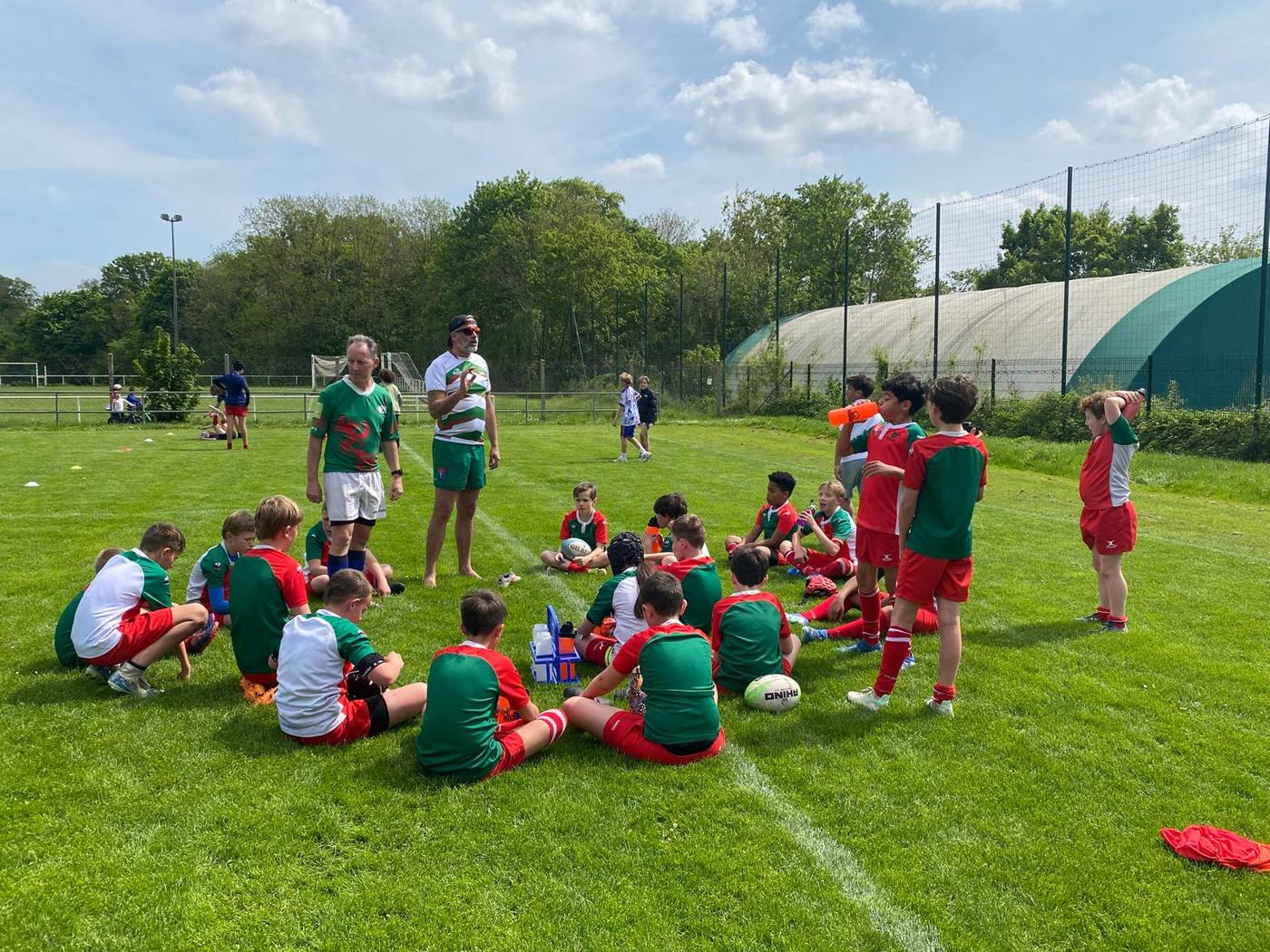 Des enfants en maillots vert et rouge assis sur l'herbe autour de leurs éducateurs, pendant un entraînement.